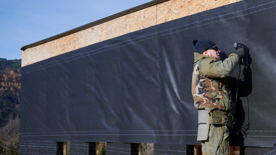 A man wearing coat, hat and sunglasses, holding an electric drill. He's installing black siding on exterior walls, using a cladding material to provide a moisture barrier
