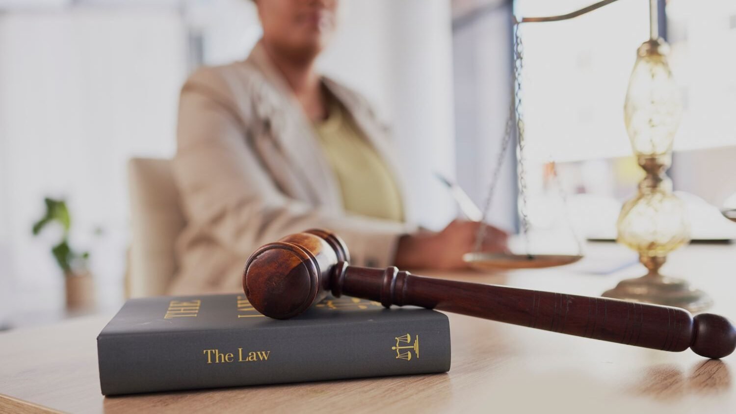 A judge in the background, out of focus. In front, there is a law book on the table, next to a judge's gavel and decorative golden scales.